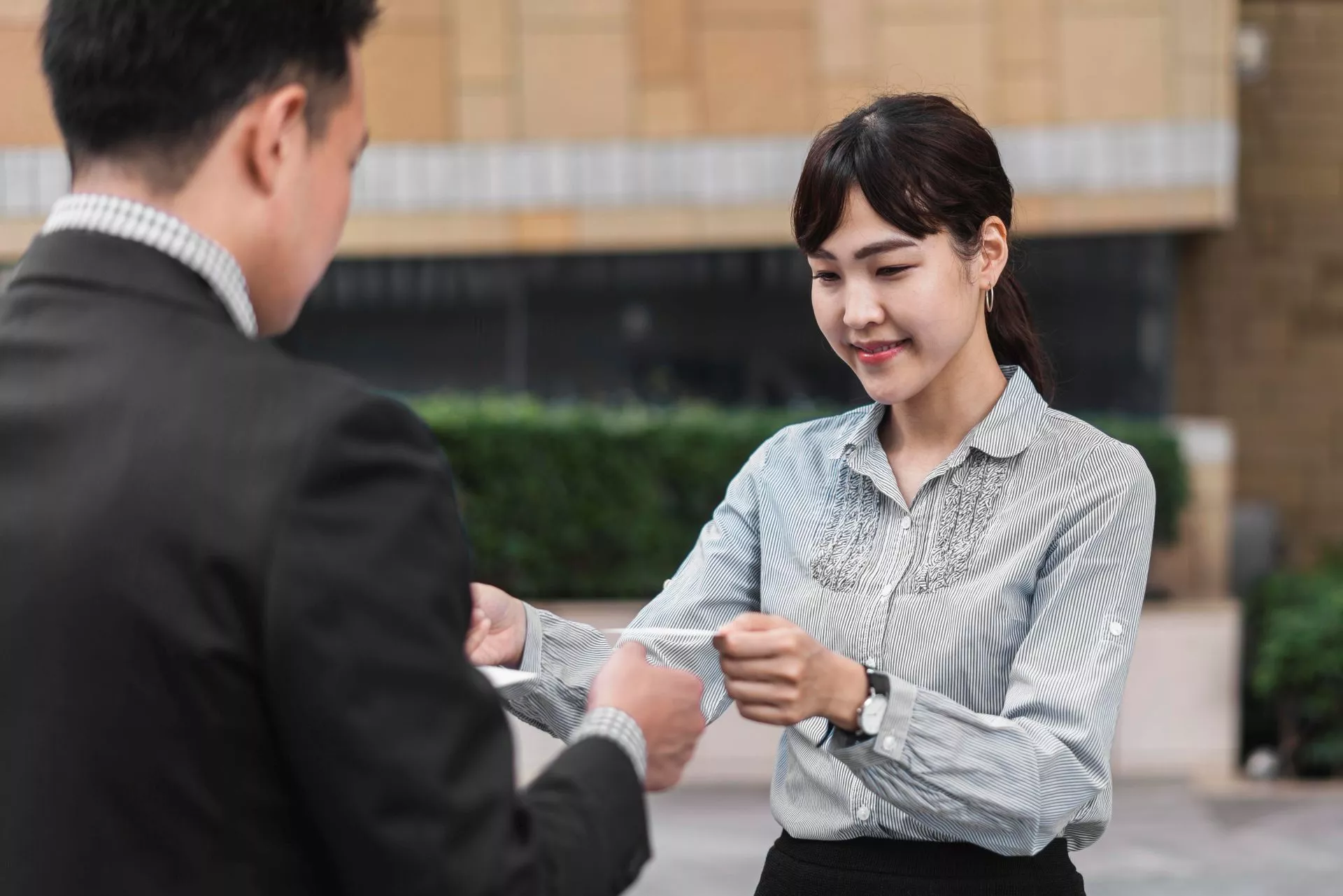 一名女性在戶外商業環境中微笑將文件遞交給男性，呈現雙方進行正式文件交付或業務往來的情境。